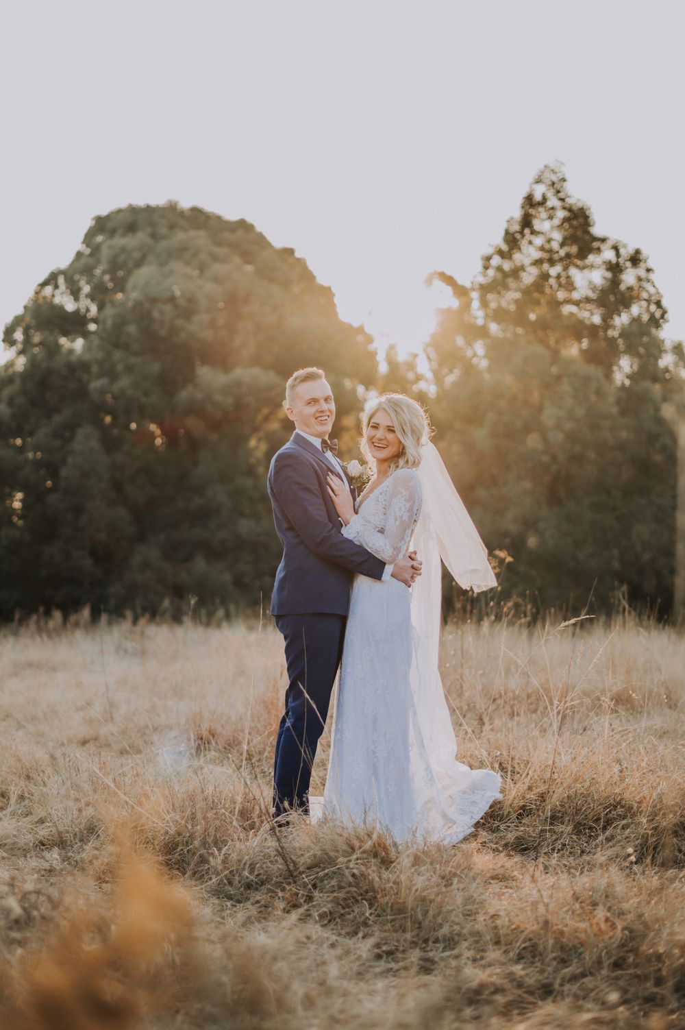 Wedding couple standing together in a field during golden hour with trees behind them.