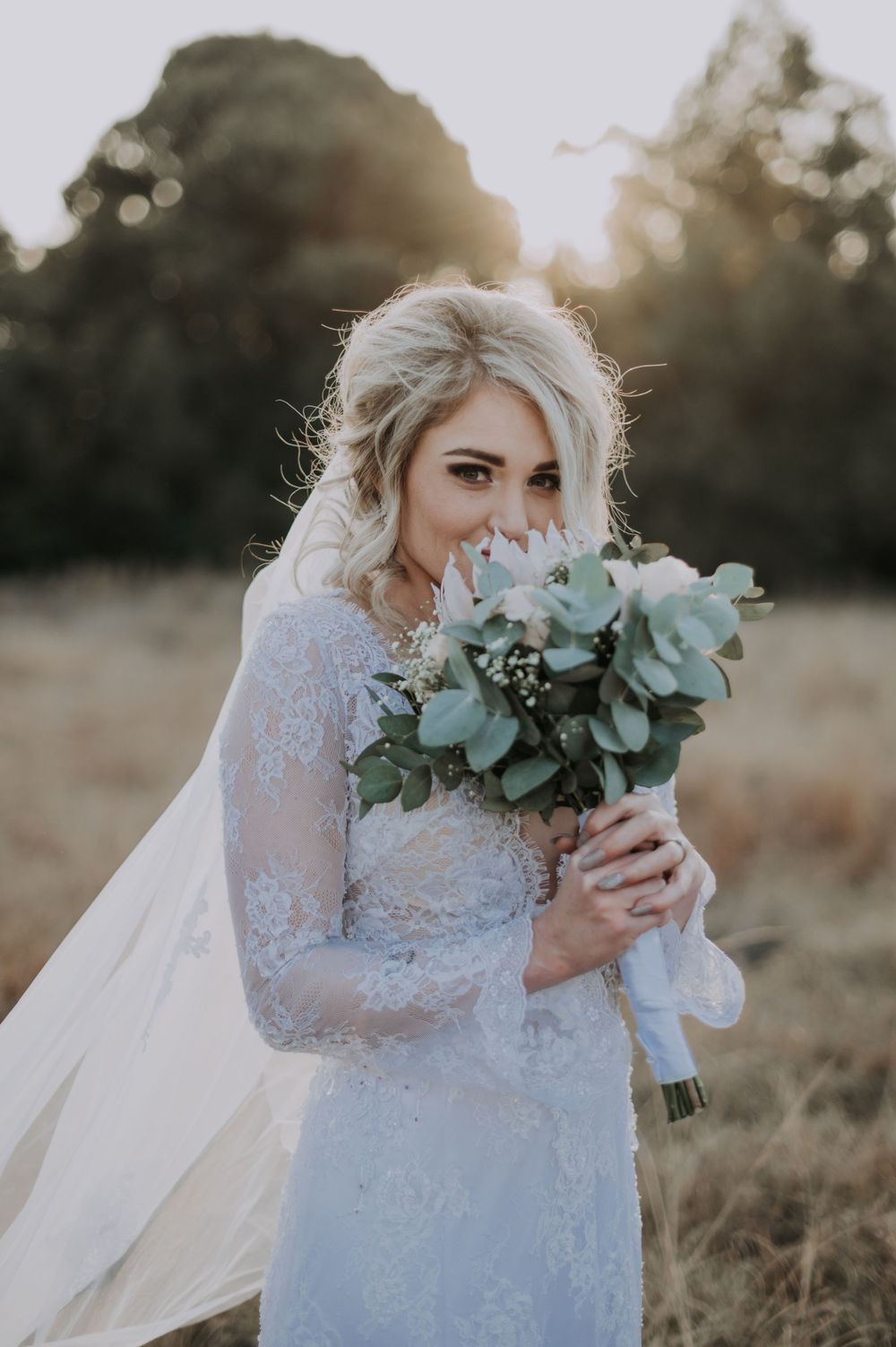 Bride holding eucalyptus bouquet while wearing long sleeve lace wedding gown at sunset.