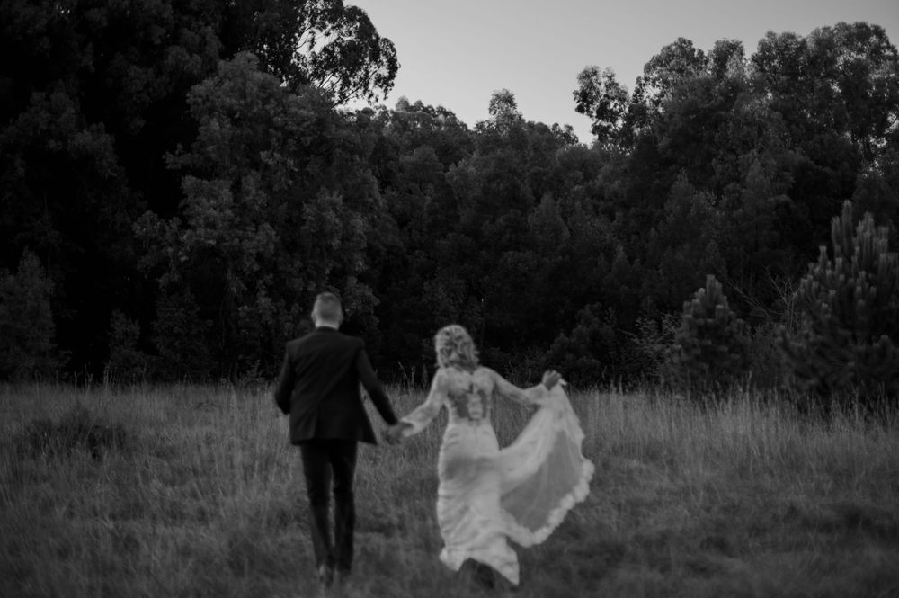 A black and white photo of newlyweds running through a field with the bride's dress flowing behind.