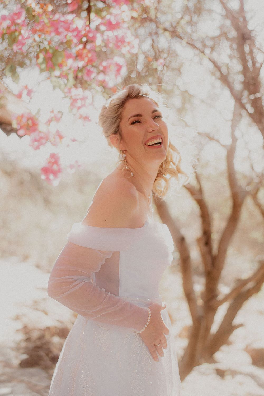 A joyful bride in an off-shoulder white dress standing under blooming pink spring trees.