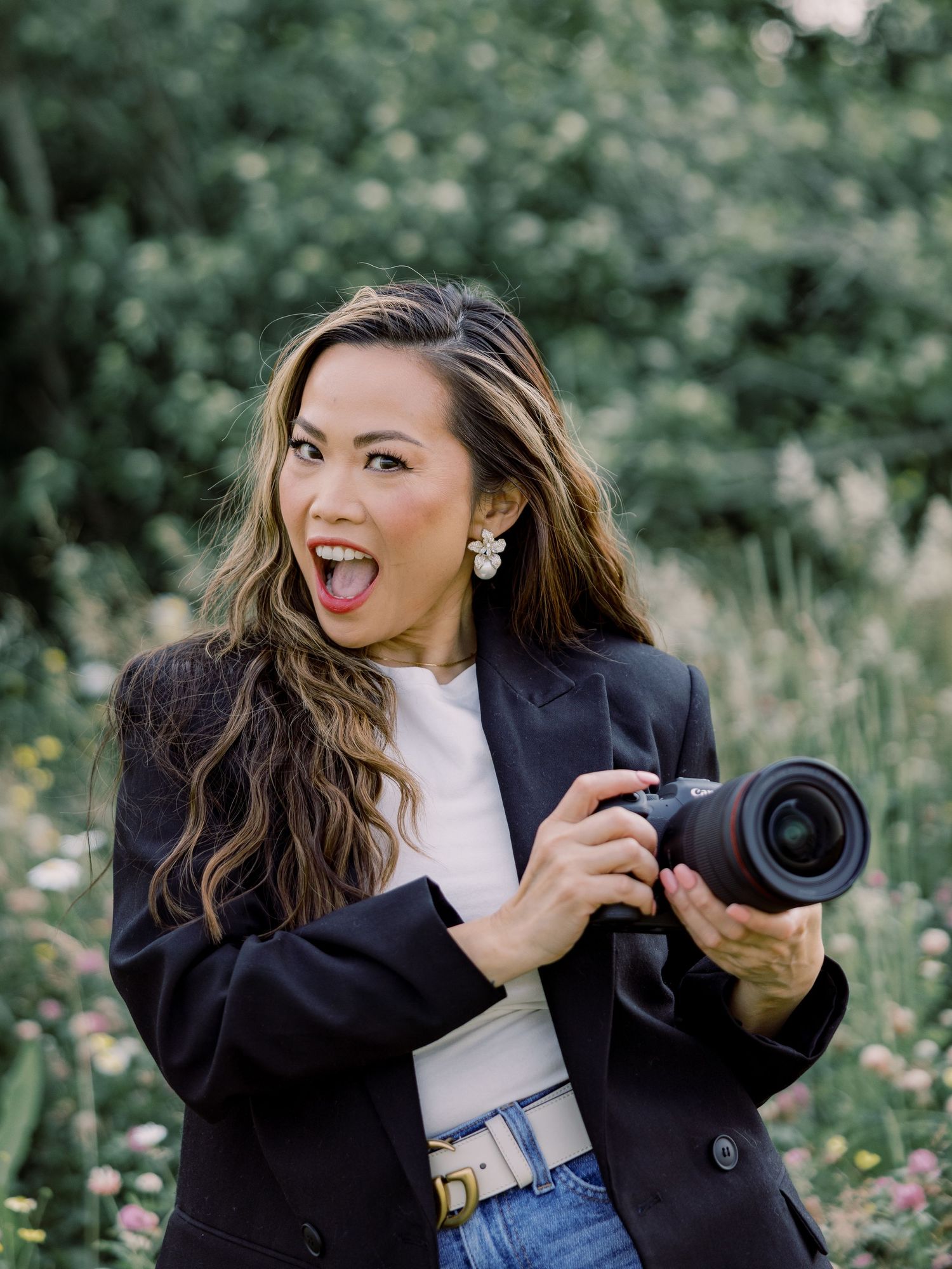 Photographer wearing a navy blazer and jeans holds a professional camera with an excited expression in a garden setting.