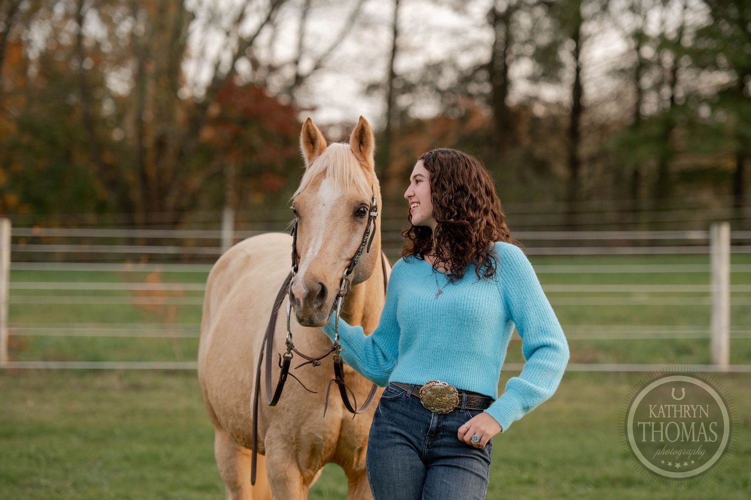 Person in bright blue sweater interacts with palomino horse in fenced paddock.