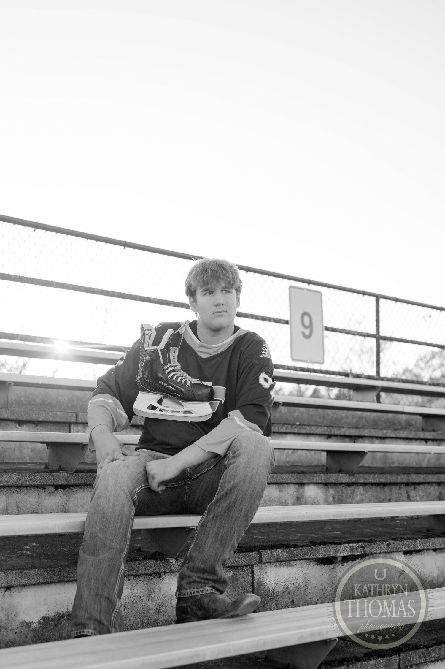 Student athlete in dark jersey sits on bleachers holding hockey skates in black and white photo.