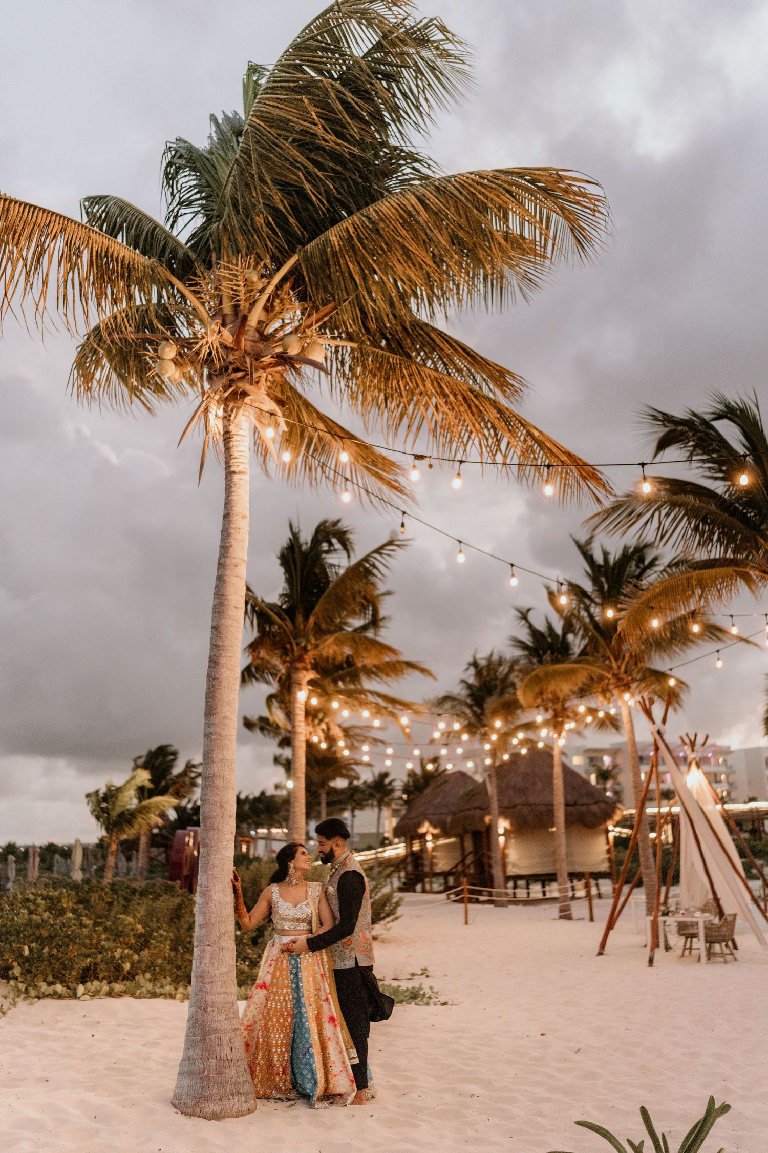 Romantic beach setting with palm trees and string lights at sunset creating a dreamy tropical atmosphere under stormy skies.