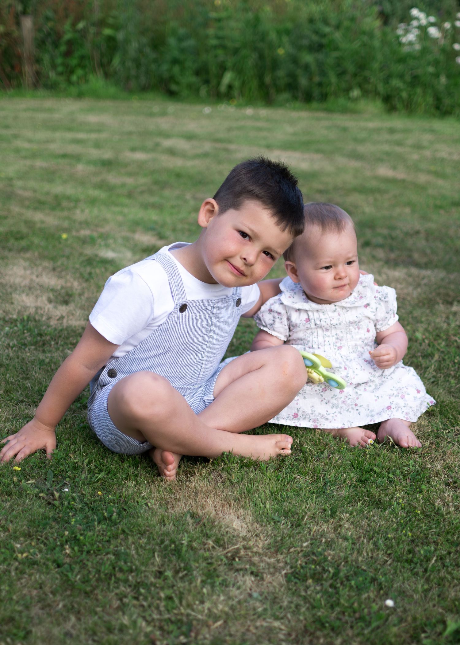 Adorable toddler boy sits on grass beside his baby sister in a sunny garden setting.