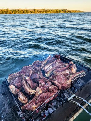 Lamb chops grilling on an open charcoal grill over a scenic river with green trees in the background.