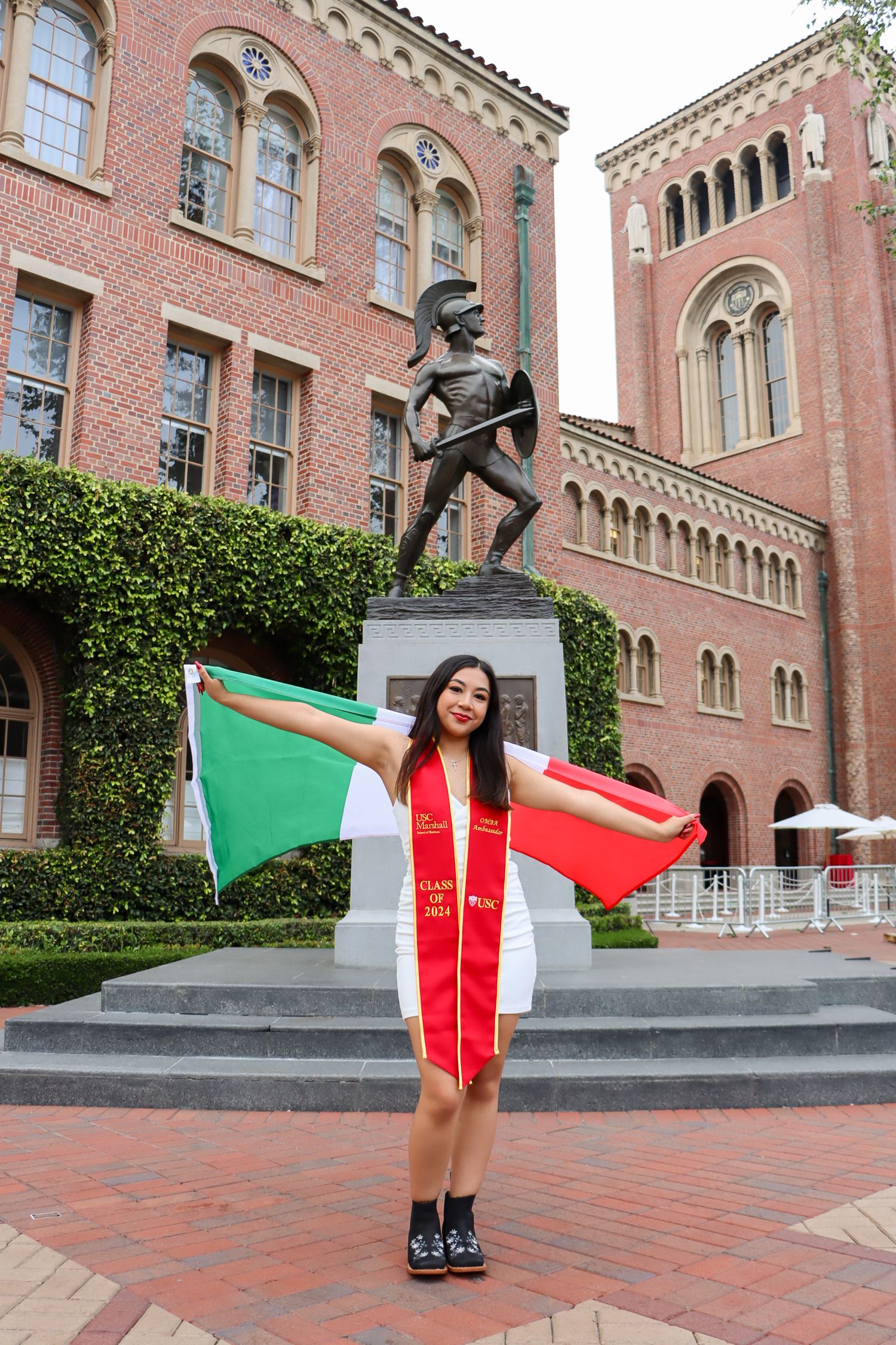 Graduate holding Italian flag at USC campus with Tommy Trojan statue in background.