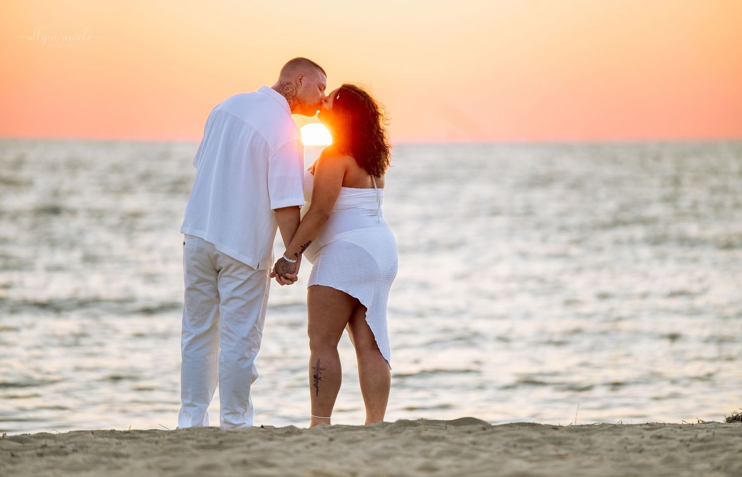 A couple in white outfits shares a romantic moment on the beach at sunset with ocean waves in the background.
