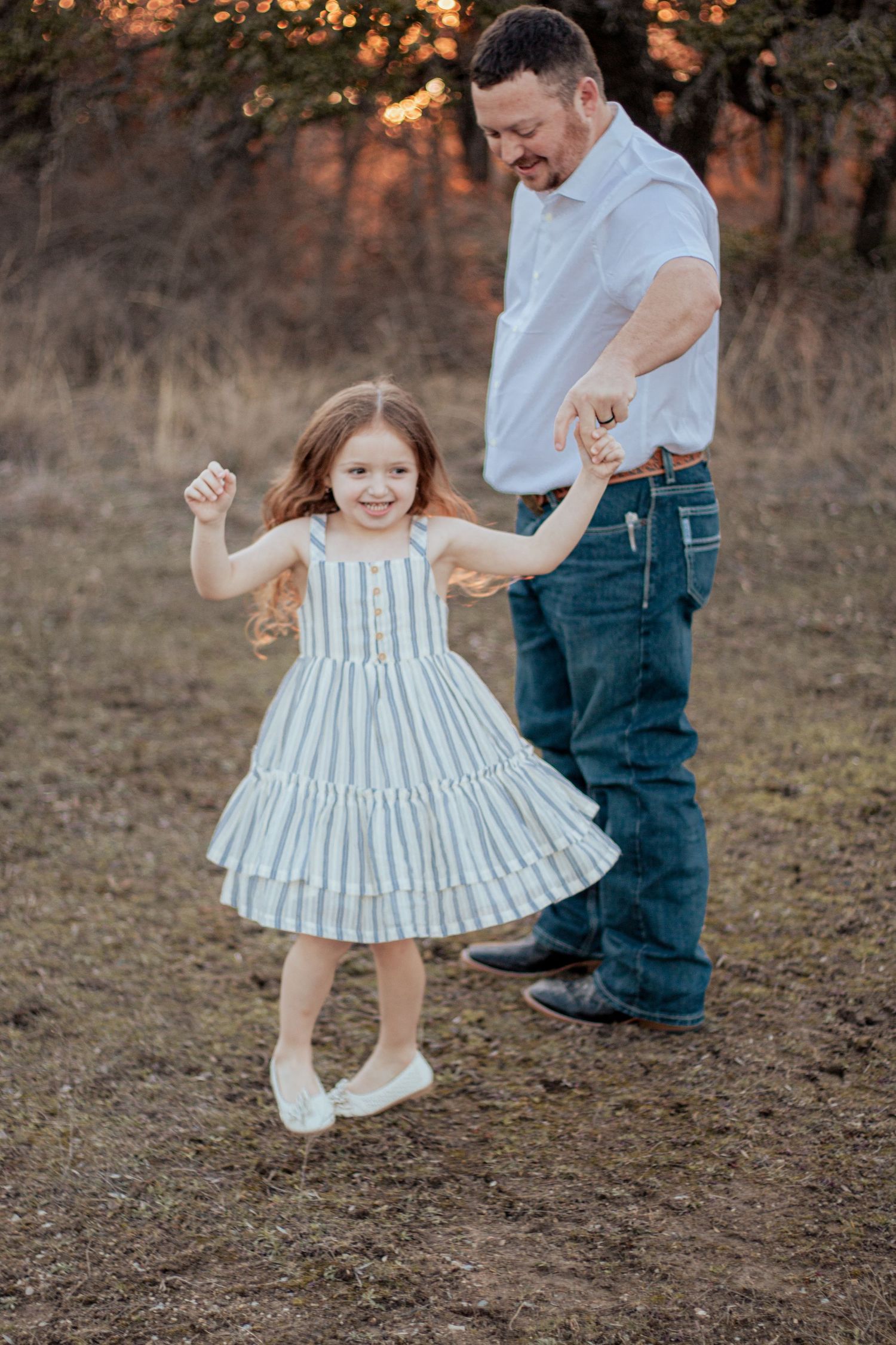 A joyful moment captured during golden hour as a child in a white tiered dress dances with a companion in a field.