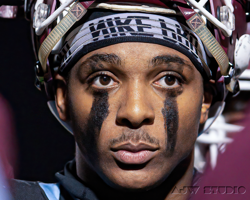 Football player wearing black and silver helmet with face paint markings against maroon background.