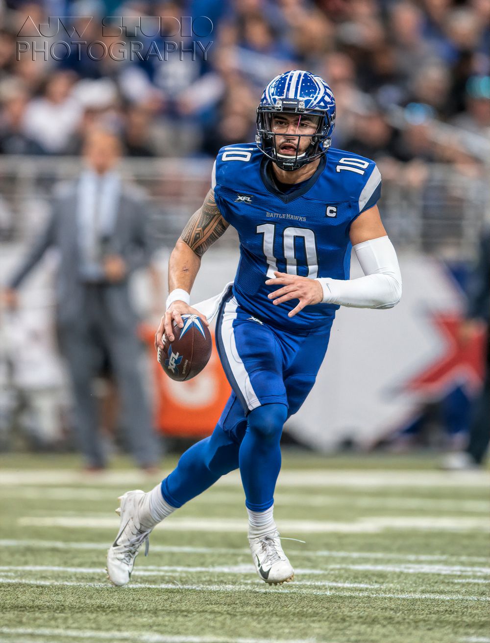 Quarterback in blue uniform runs with football during indoor stadium game.