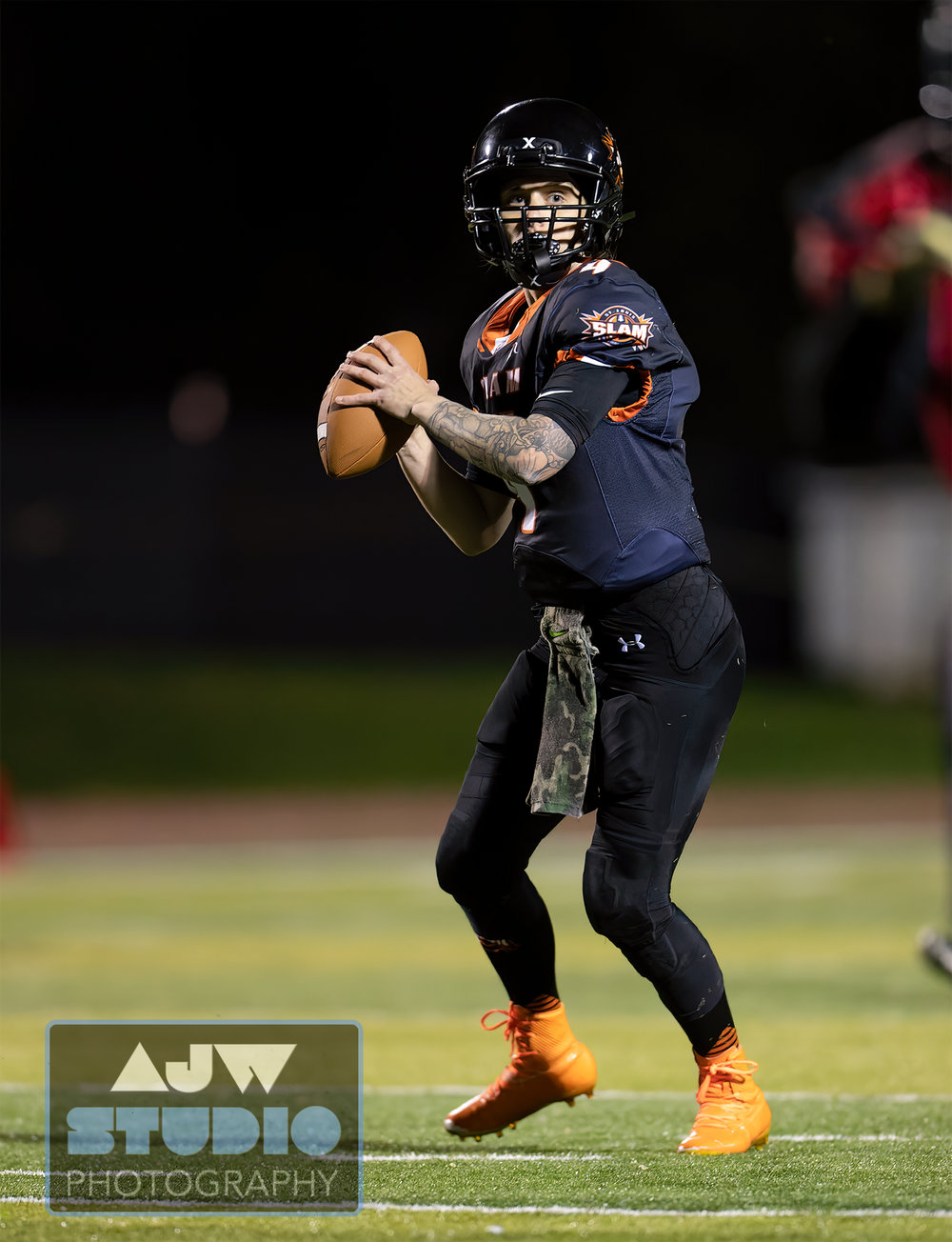 Quarterback in navy uniform sets up to throw during nighttime football game.