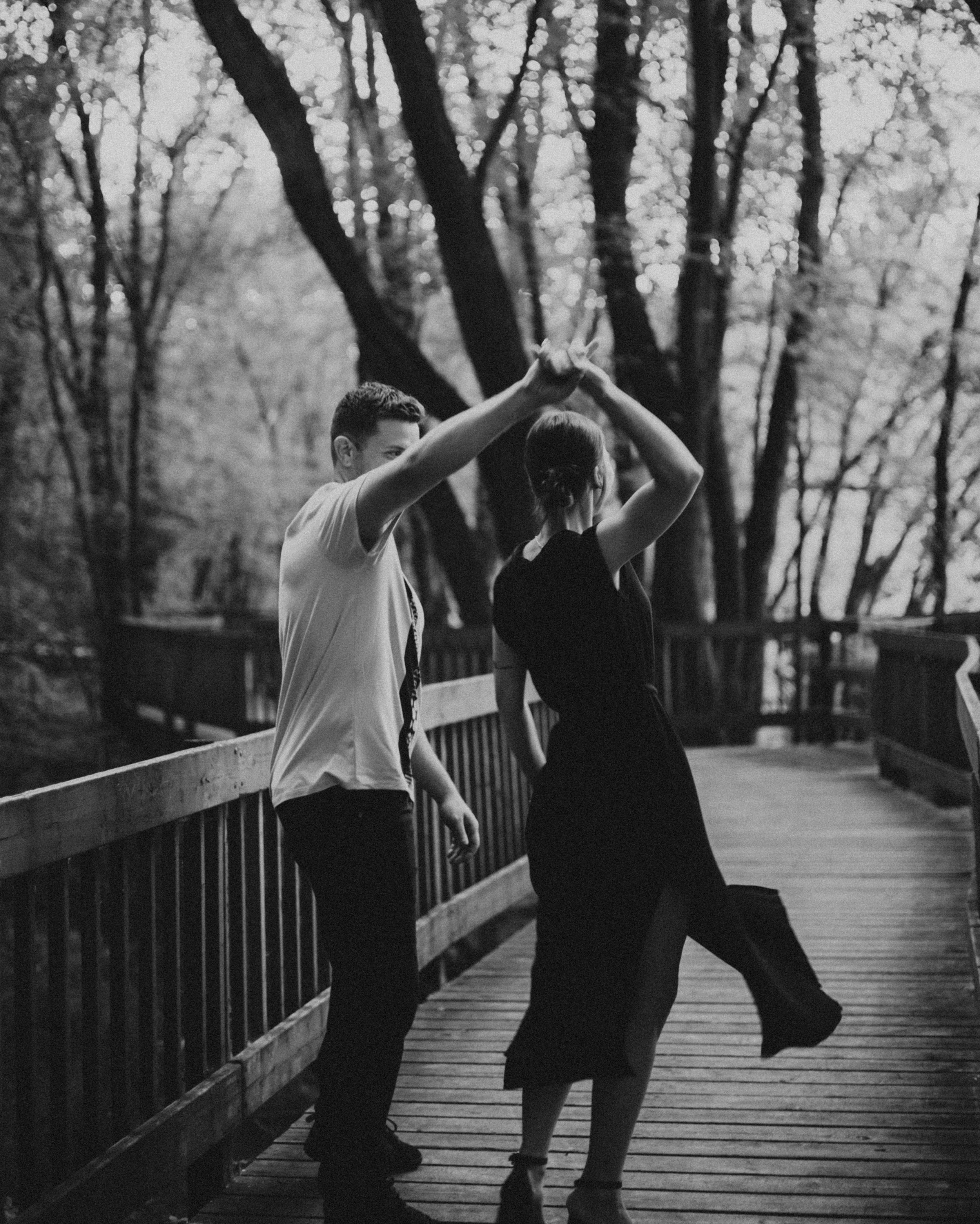 Black and white photo of couple dancing on wooden bridge surrounded by trees.