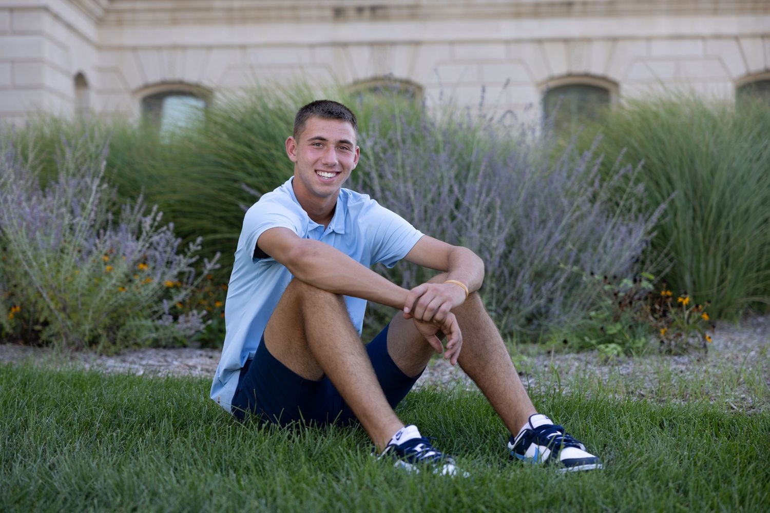 A person in a light blue t-shirt and shorts sits casually on grass near decorative lavender plants outside a building.