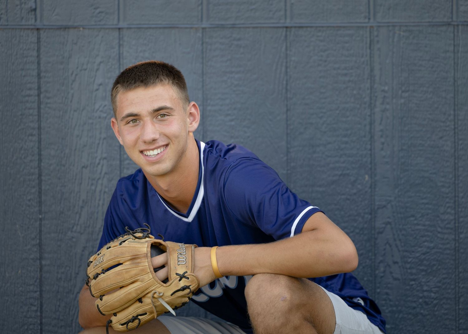 Baseball player in blue uniform smiles while posing with glove against dark gray wall.