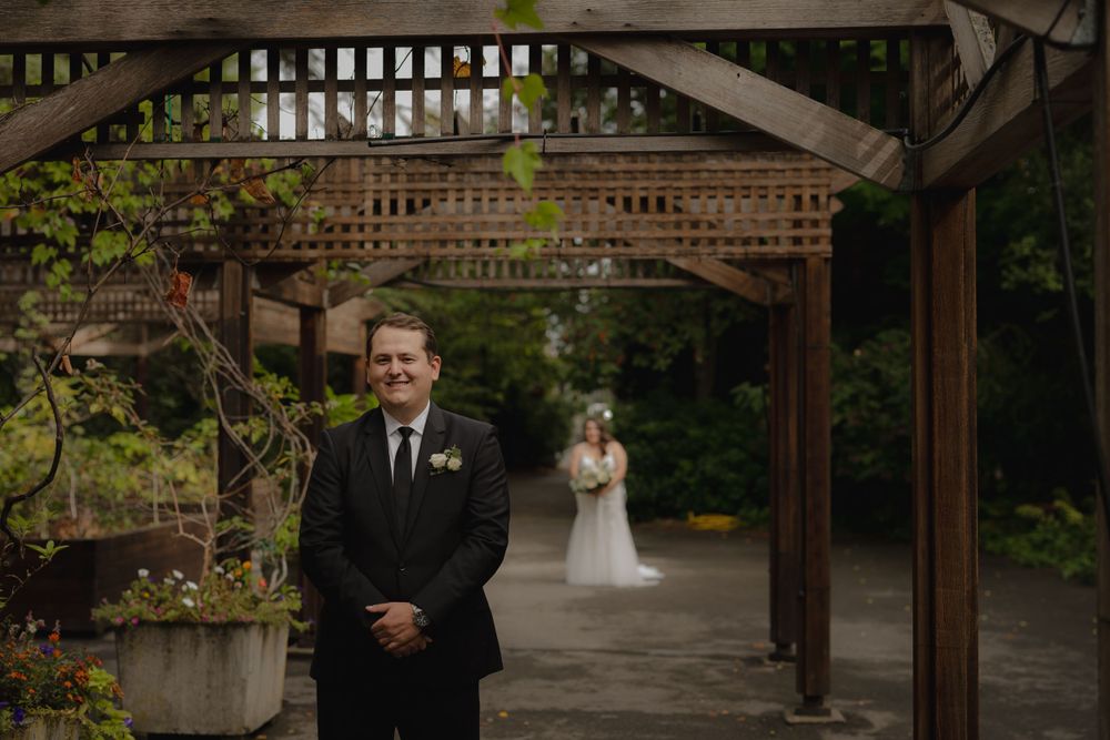 First look wedding moment captured under rustic wooden pergola with lush greenery.