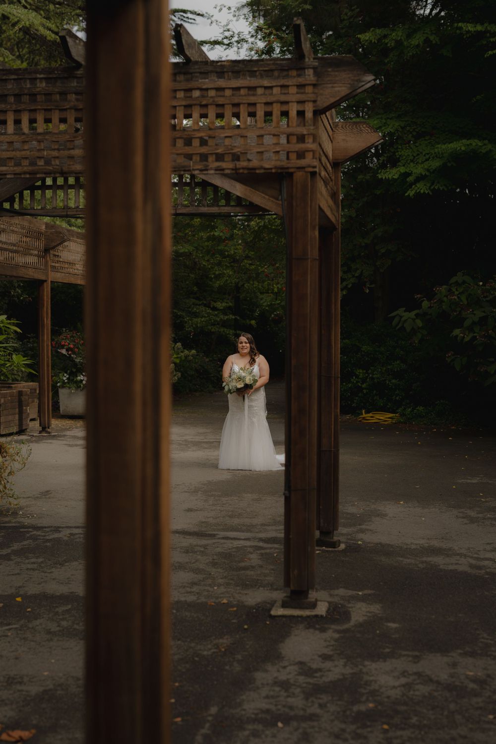 Bride in white dress stands alone under wooden pergola in garden setting.