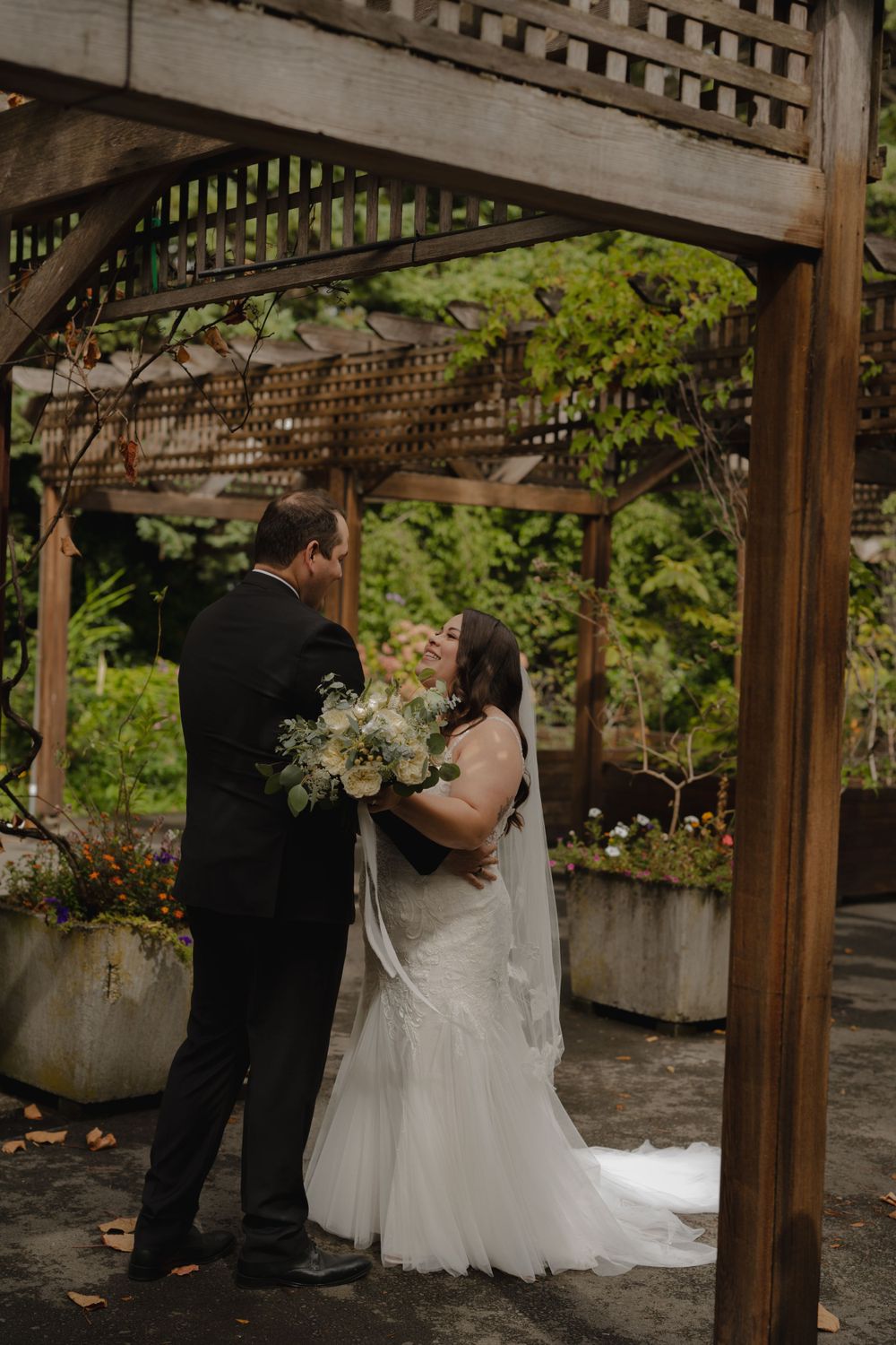 Intimate wedding moment captured under wooden pergola with garden backdrop.