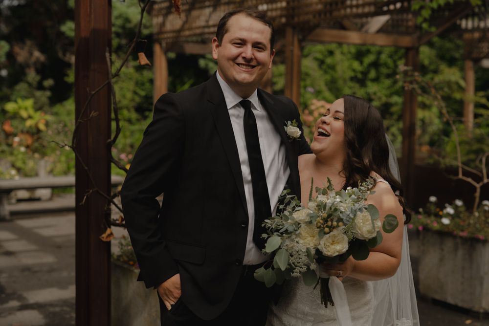A joyful couple shares a candid moment under a wooden pergola during their wedding celebration.