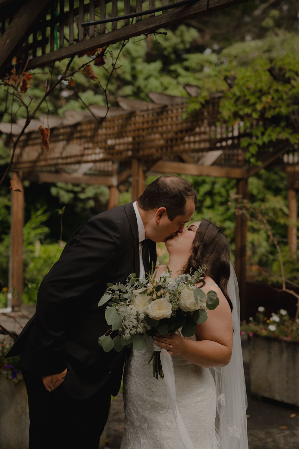 A romantic moment captured between newlyweds kissing under a garden trellis with a beautiful bouquet.