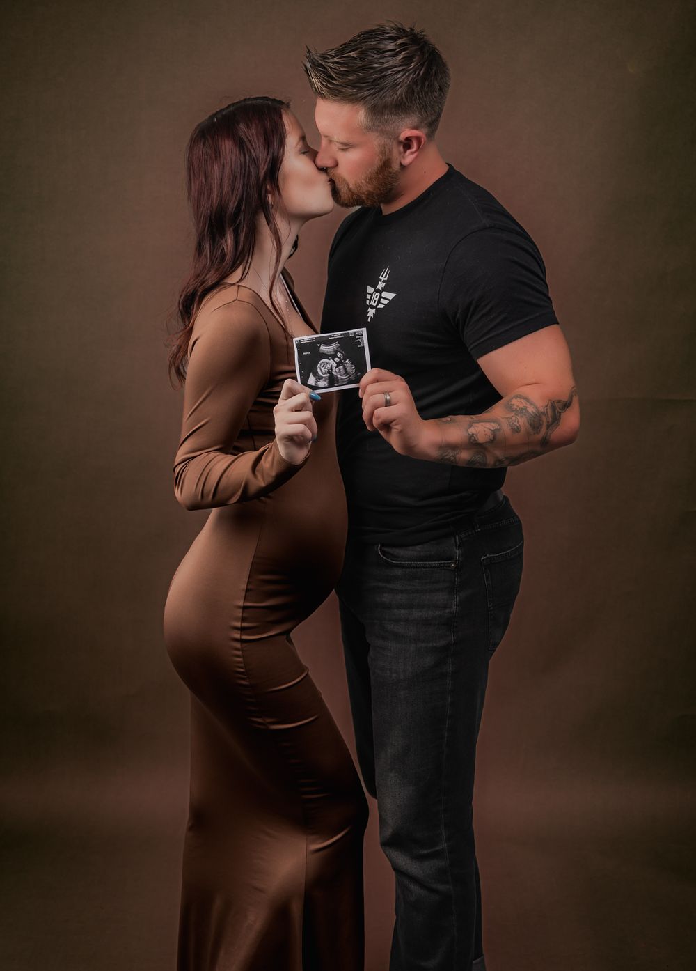Romantic maternity photo of couple in brown tones sharing intimate moment while holding ultrasound image.