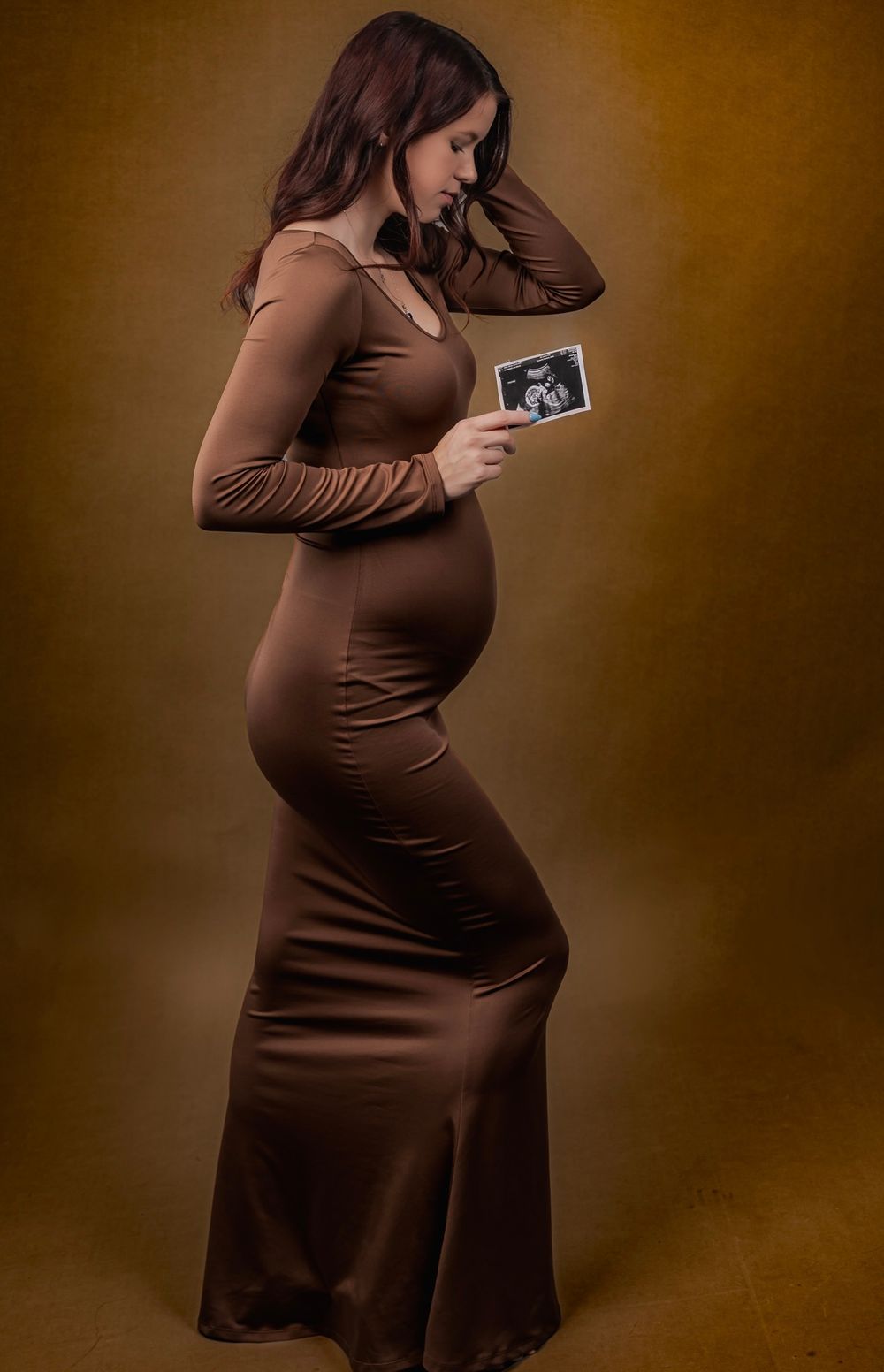 Maternity photo of woman in brown dress holding ultrasound photo against warm backdrop.