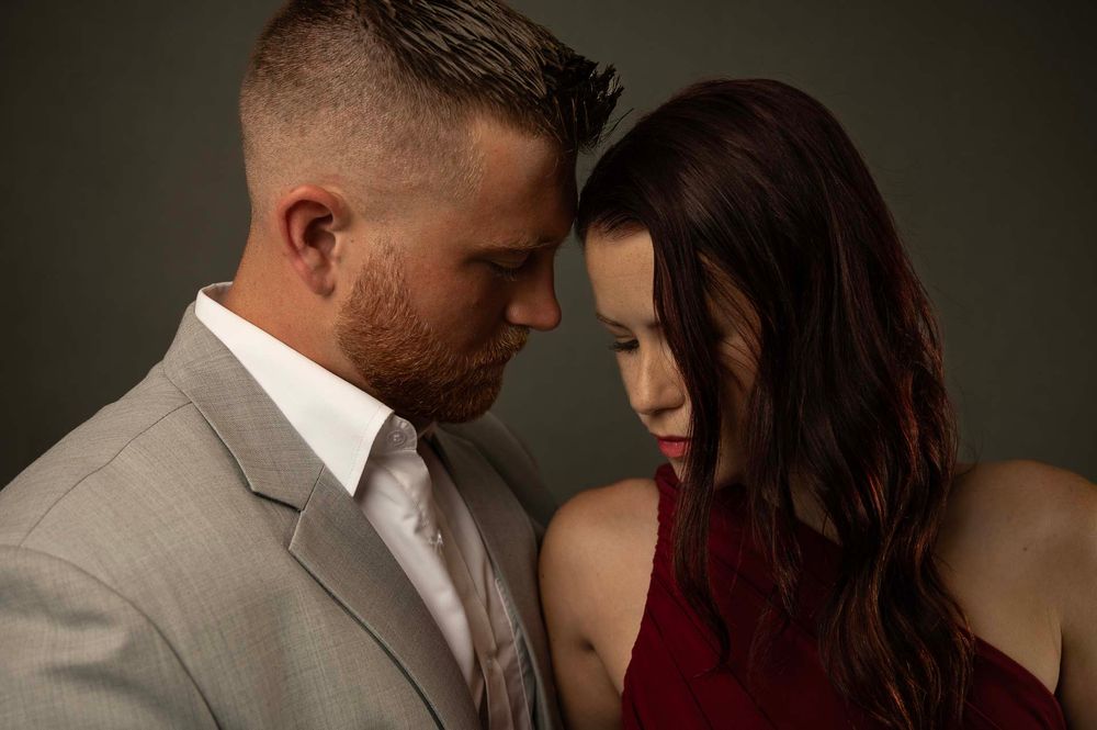 A couple in formal attire share an intimate moment against a dark backdrop, with him in grey suit and her in red dress.