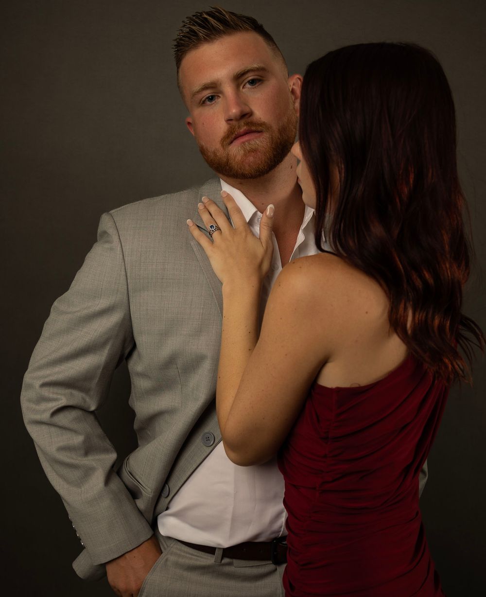 A romantic couple embraces in an intimate portrait, with a woman in red touching the person wearing a gray suit.