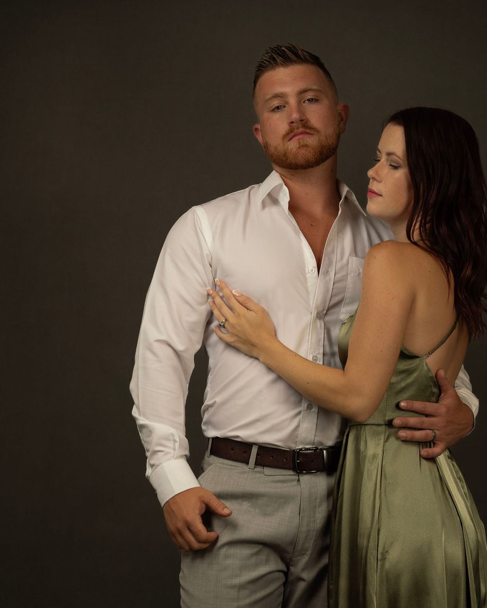 A romantic couple embraces in studio lighting, with the woman wearing a sage green dress and the man in a white dress shirt.