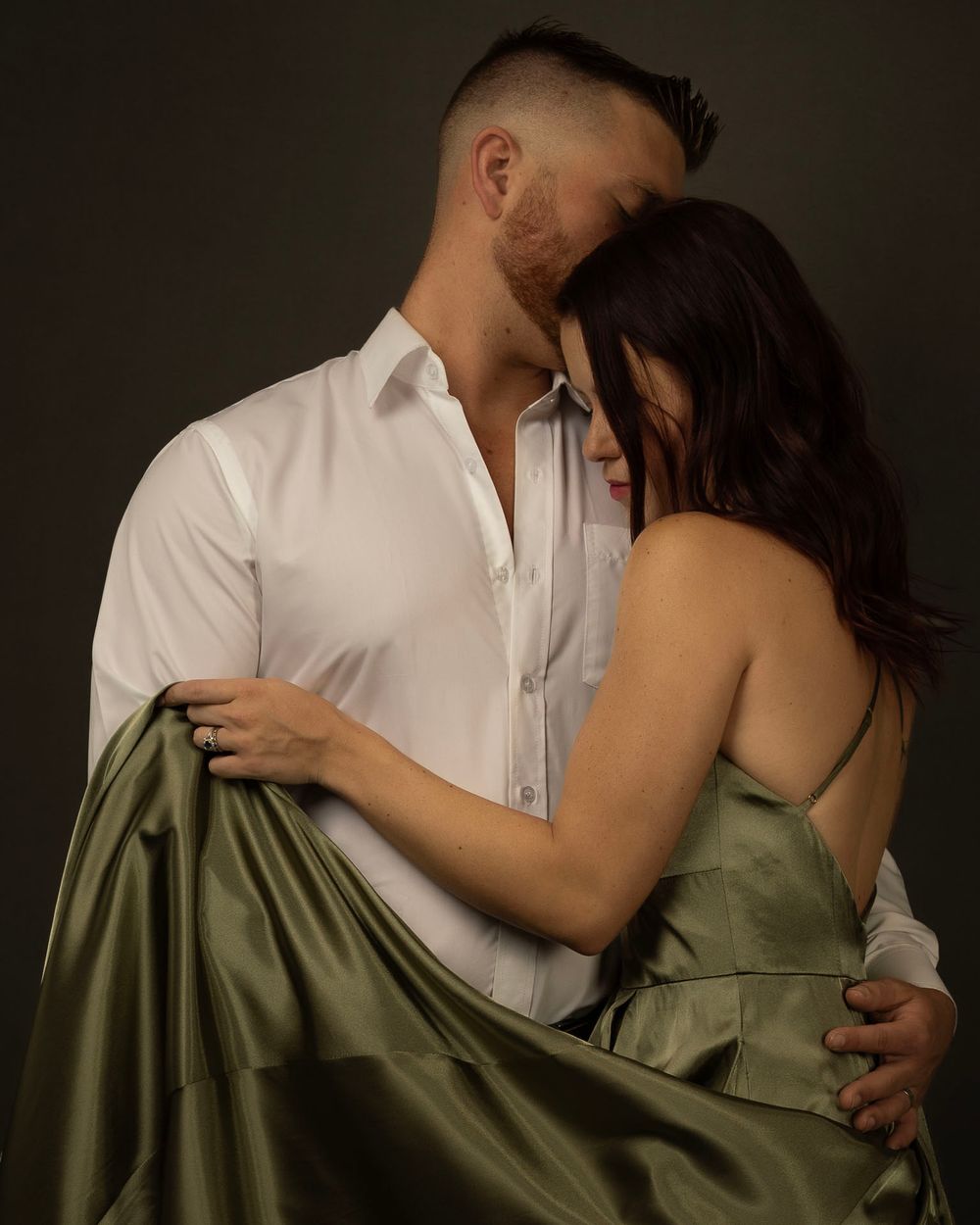 Intimate couple embraces in dramatic lighting with figure in white shirt and other in flowing sage green dress.