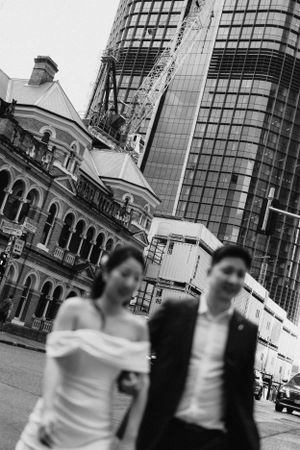 A black and white urban wedding photo showing a couple walking past historic architecture and modern buildings in the city.