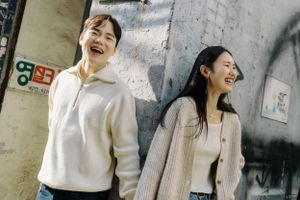 Two people in cream colored sweaters laugh while standing against a gray concrete wall outdoors.
