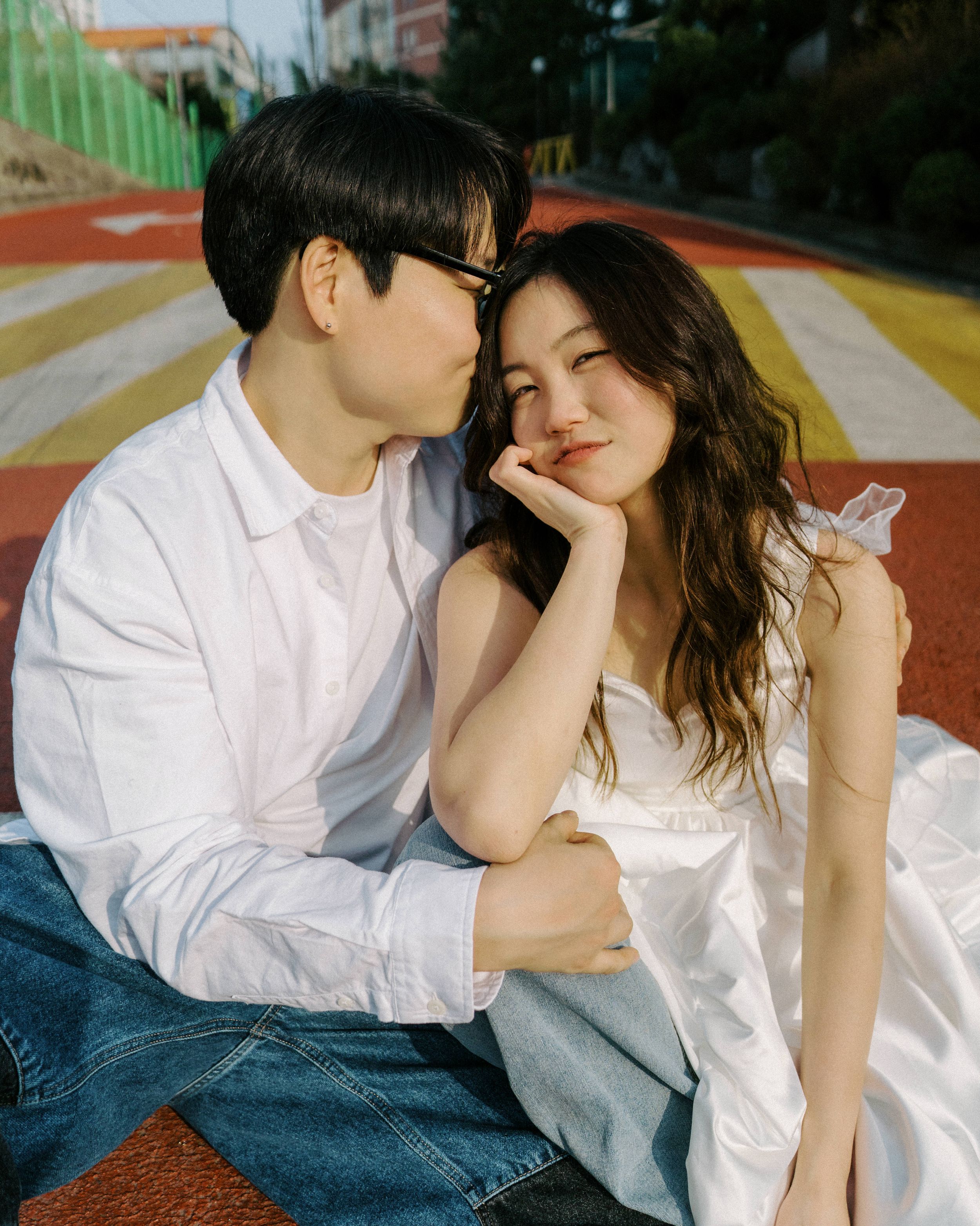 A couple in white outfits sits intimately on a colorful striped outdoor track sharing a tender moment together.