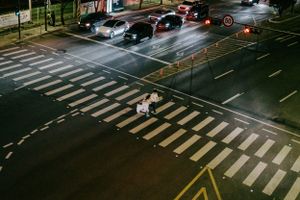 Two pedestrians cross a well-lit zebra crossing at night on a multi-lane road while cars wait at the intersection.