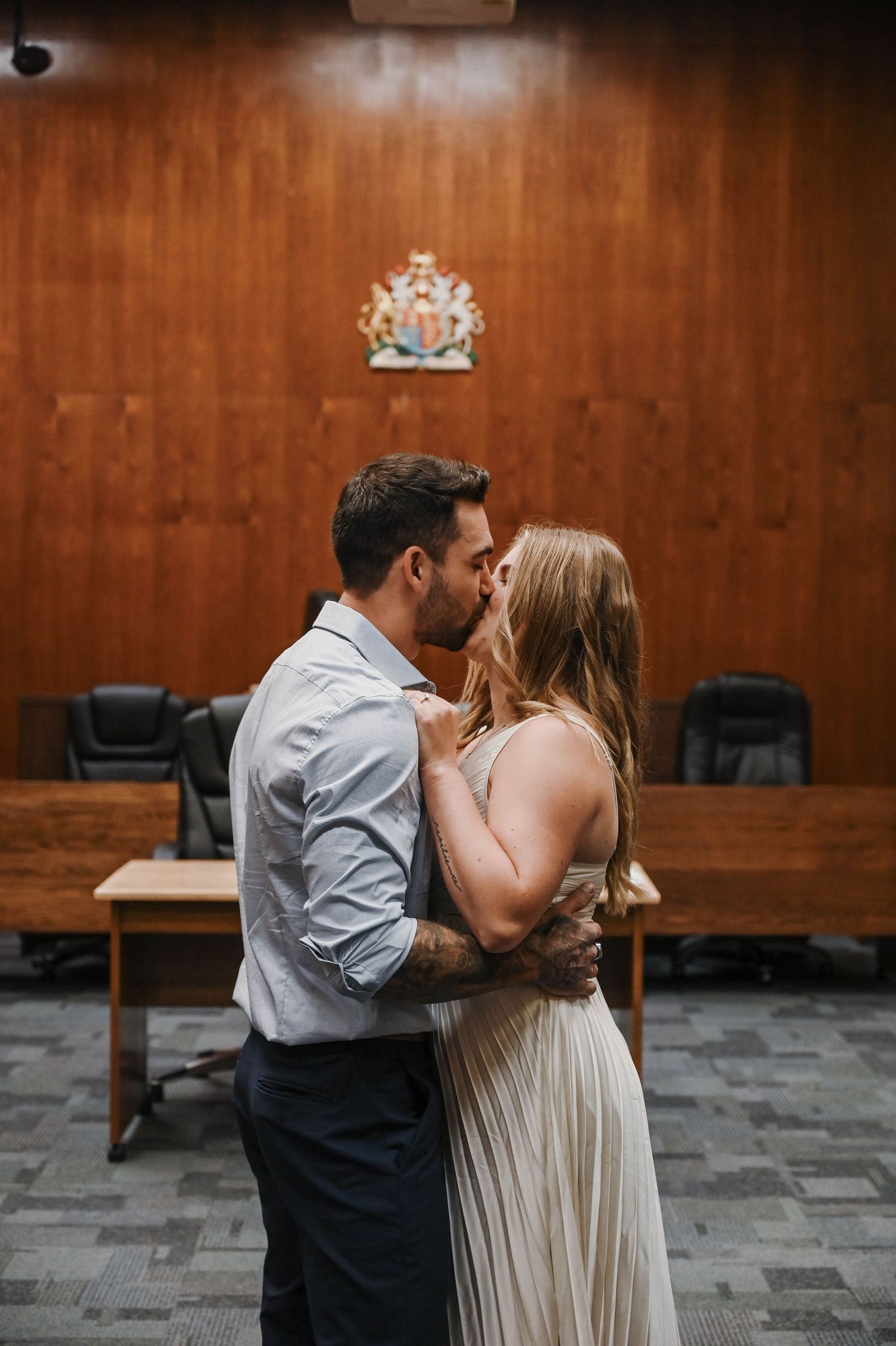 A couple shares an intimate kiss in a courthouse room with wood paneled walls and a coat of arms mounted above.