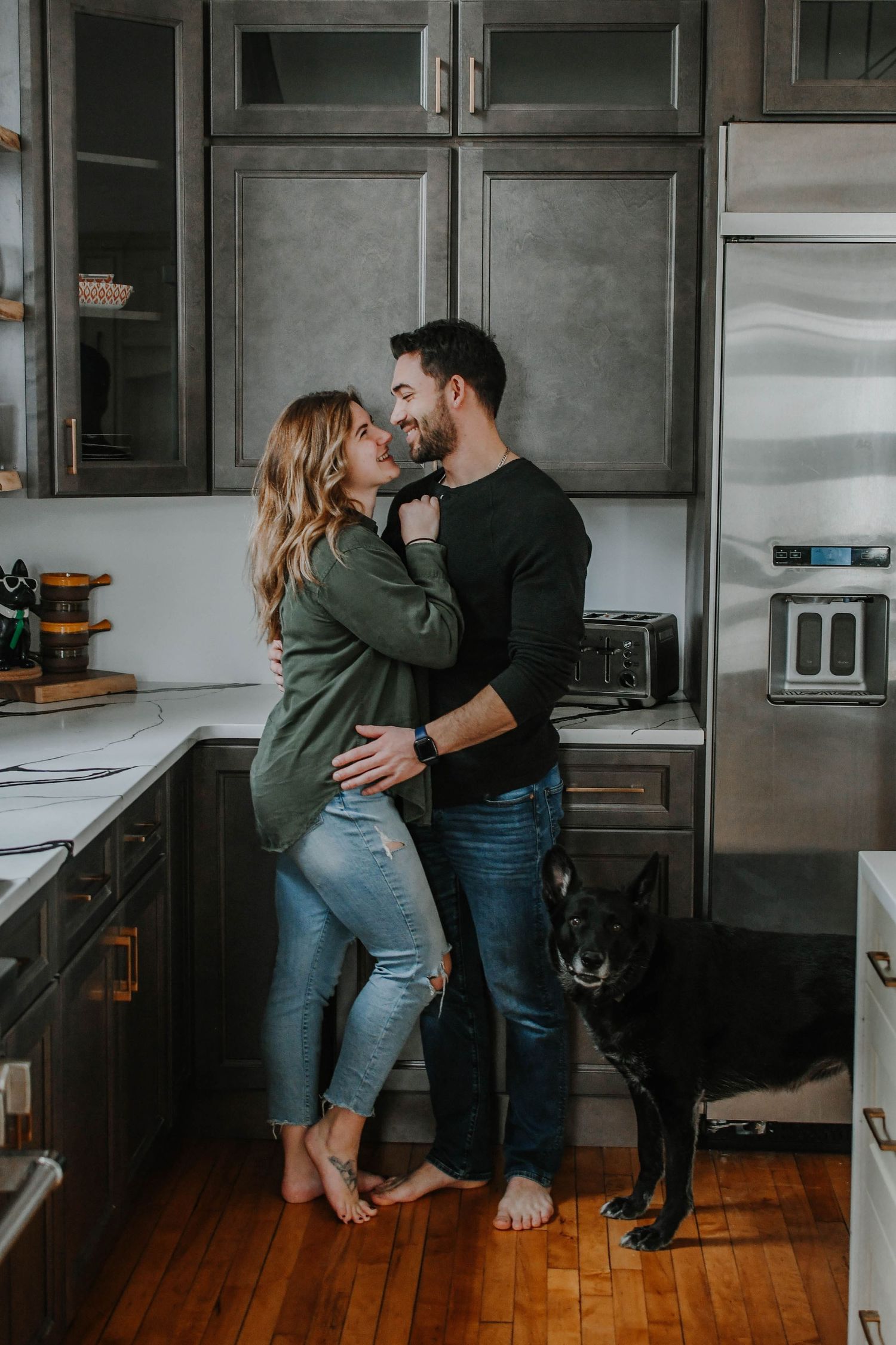 Couple embraces in cozy kitchen with hardwood floors while black dog watches nearby.