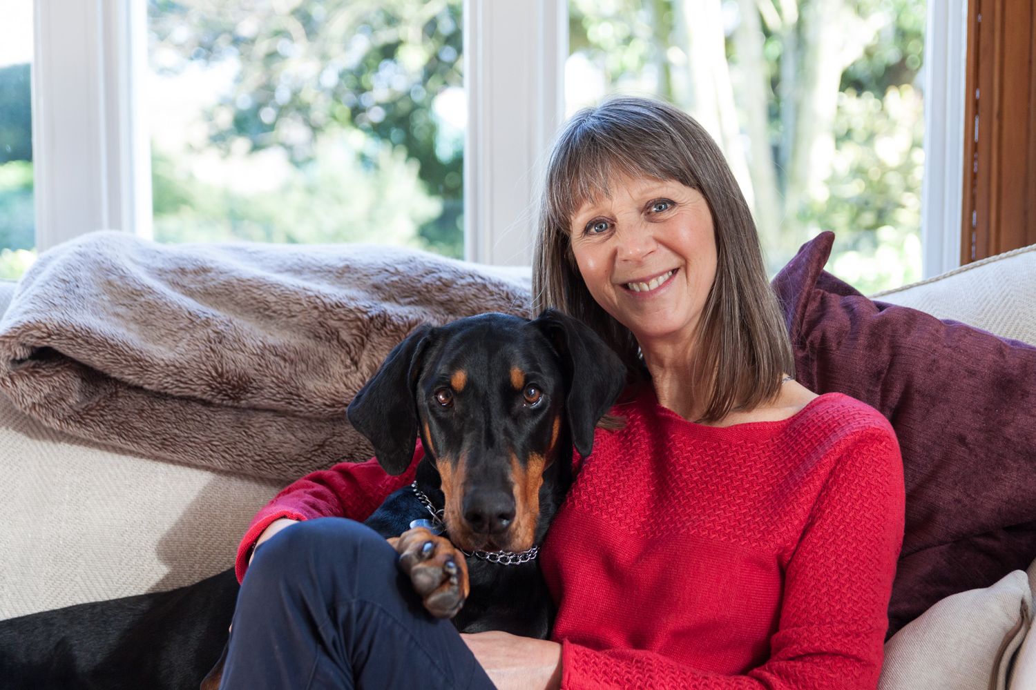 A lady in a red jumper cuddles up on the sofa with her Doberman. North Wales Portrait Photographer