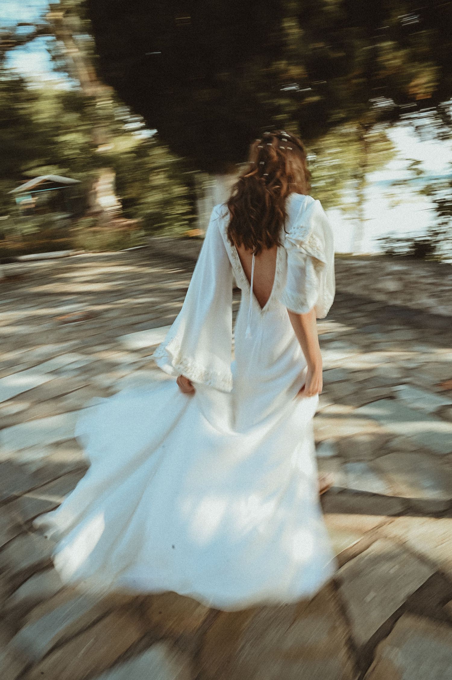 A flowing white dress with a deep V-back moves gracefully on a sunlit stone path through dappled shadows.