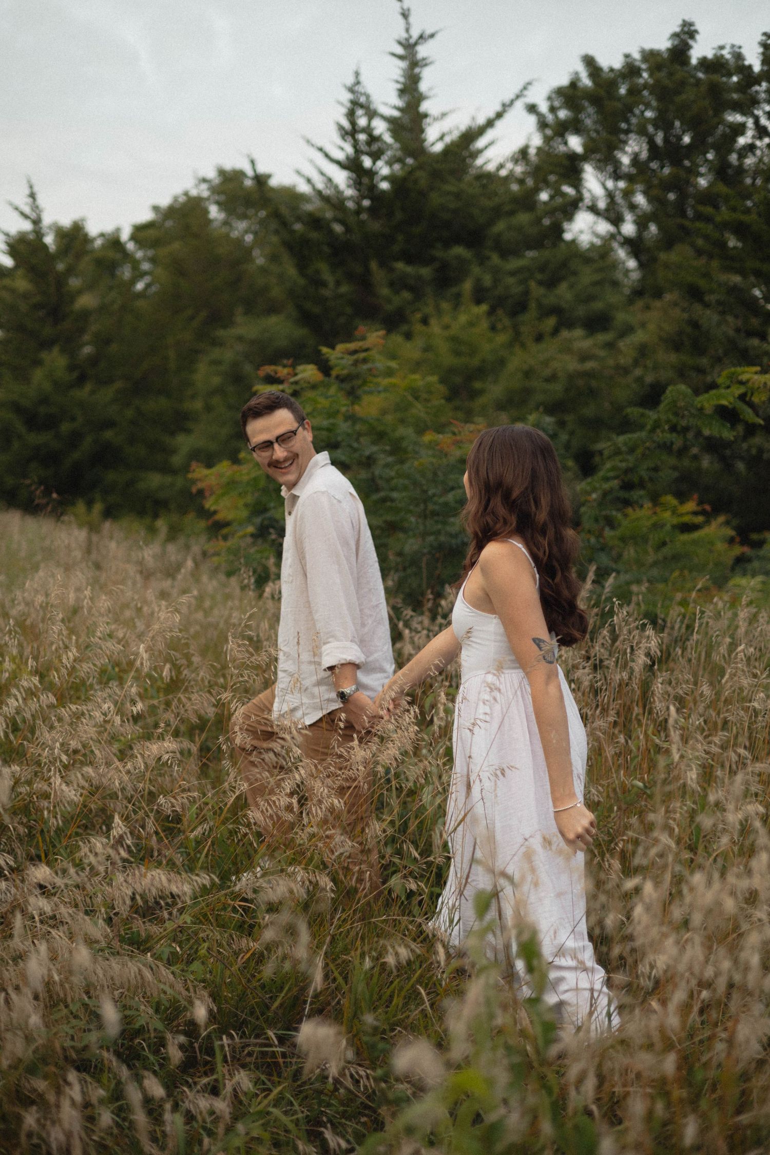 Couple holding hands while walking through tall grass and evergreen trees in natural outdoor setting.
