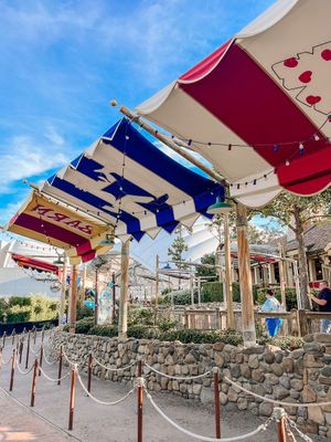 Decorative red, white, and blue awnings with hearts adorn a stone wall structure at a Disney theme park entrance.
