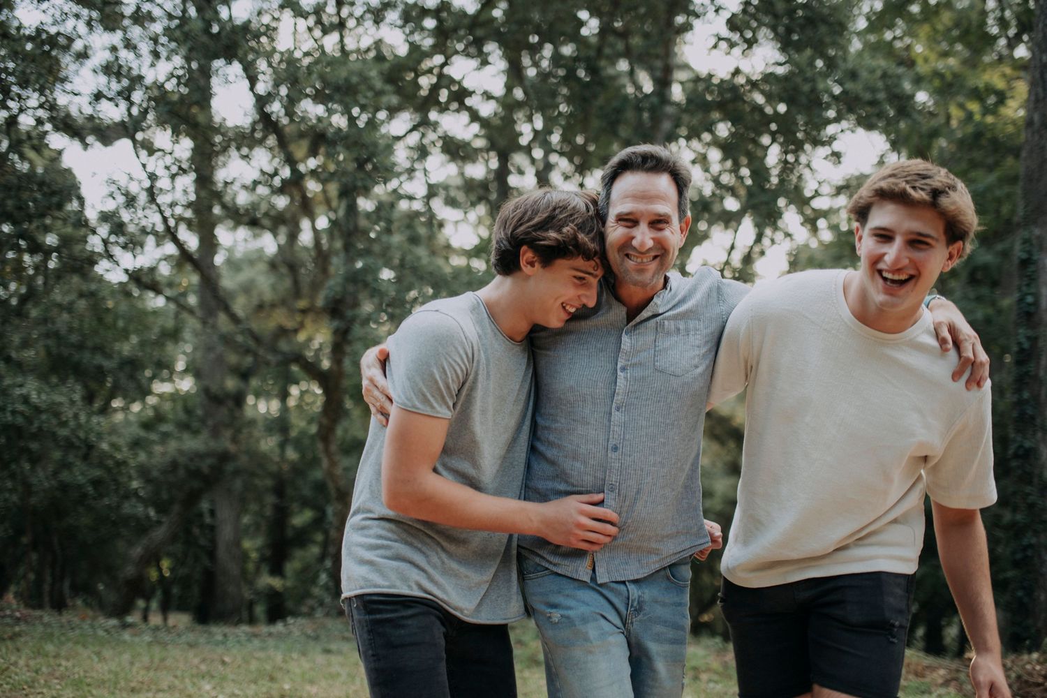 Three people sharing a candid laughing moment together outdoors with trees in the background.