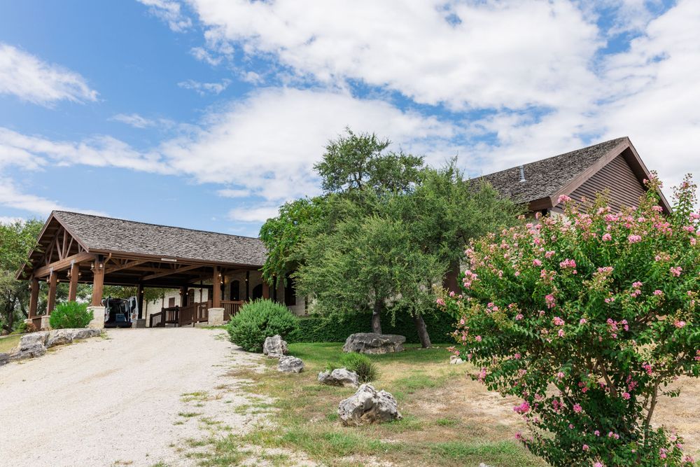 Rural lodge buildings with wooden architecture and a gravel driveway surrounded by green trees and blooming shrubs.