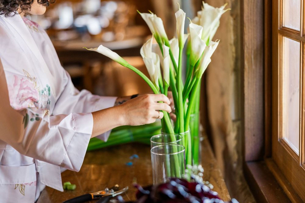 Hands placing fresh white calla lilies in a glass vase by a rustic window.