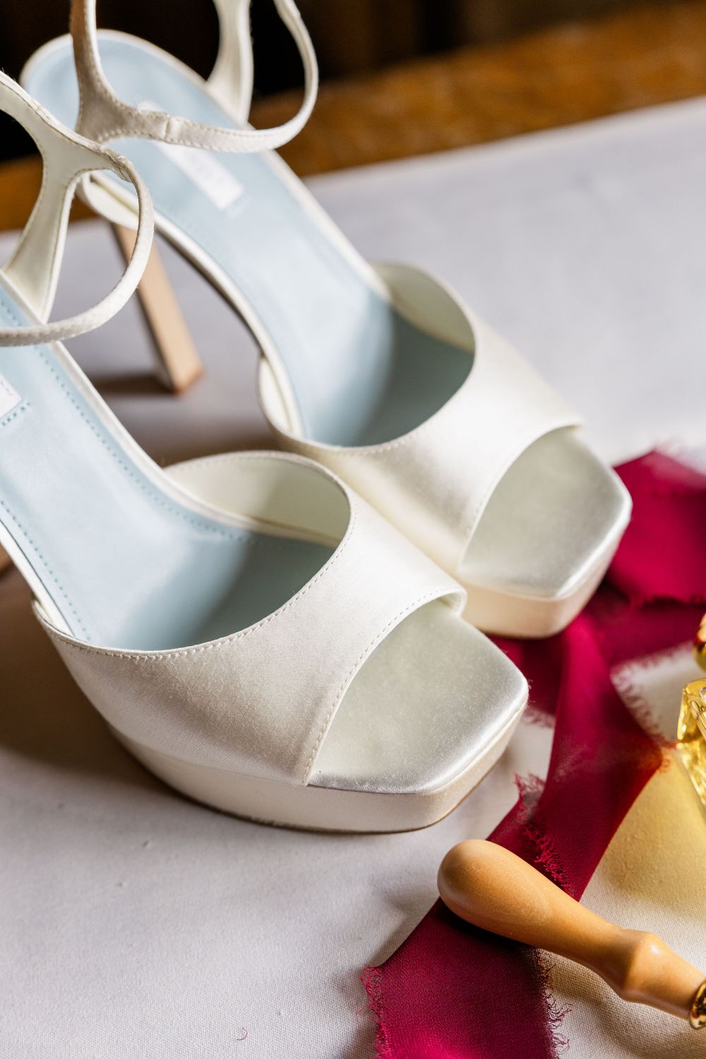 Elegant white satin platform wedding shoes with ankle straps displayed next to a burgundy ribbon.