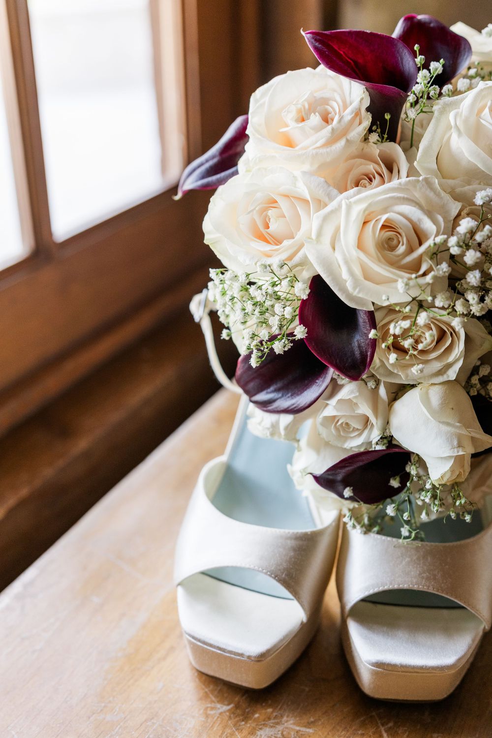 A bridal bouquet with white roses and deep burgundy calla lilies sits next to white wedding heels near a window.