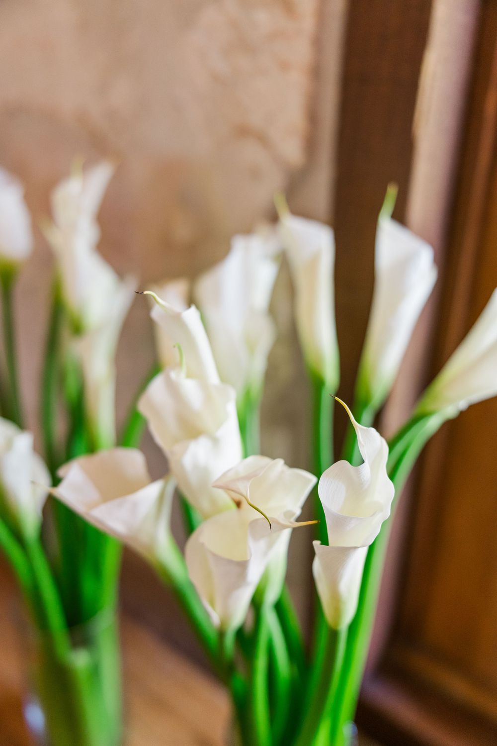 White calla lily flowers with green stems arranged in soft focus against a warm beige background.