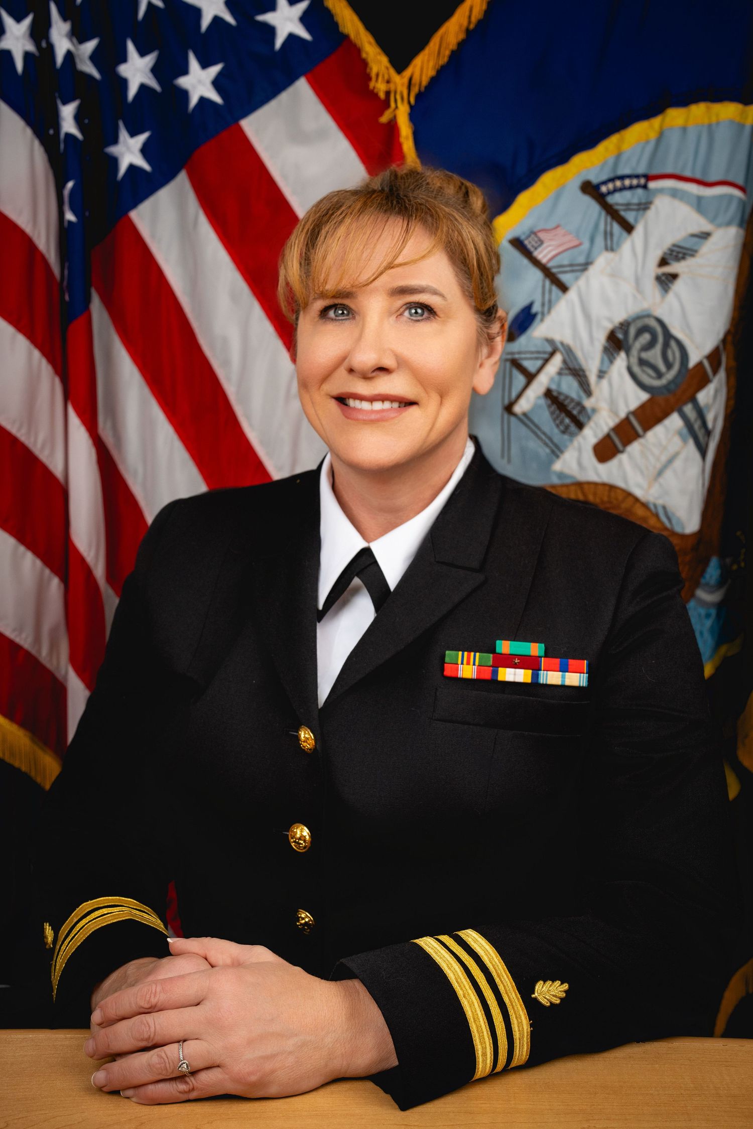 A naval officer in dress uniform poses for an official portrait with the American flag and Navy emblem in the background.