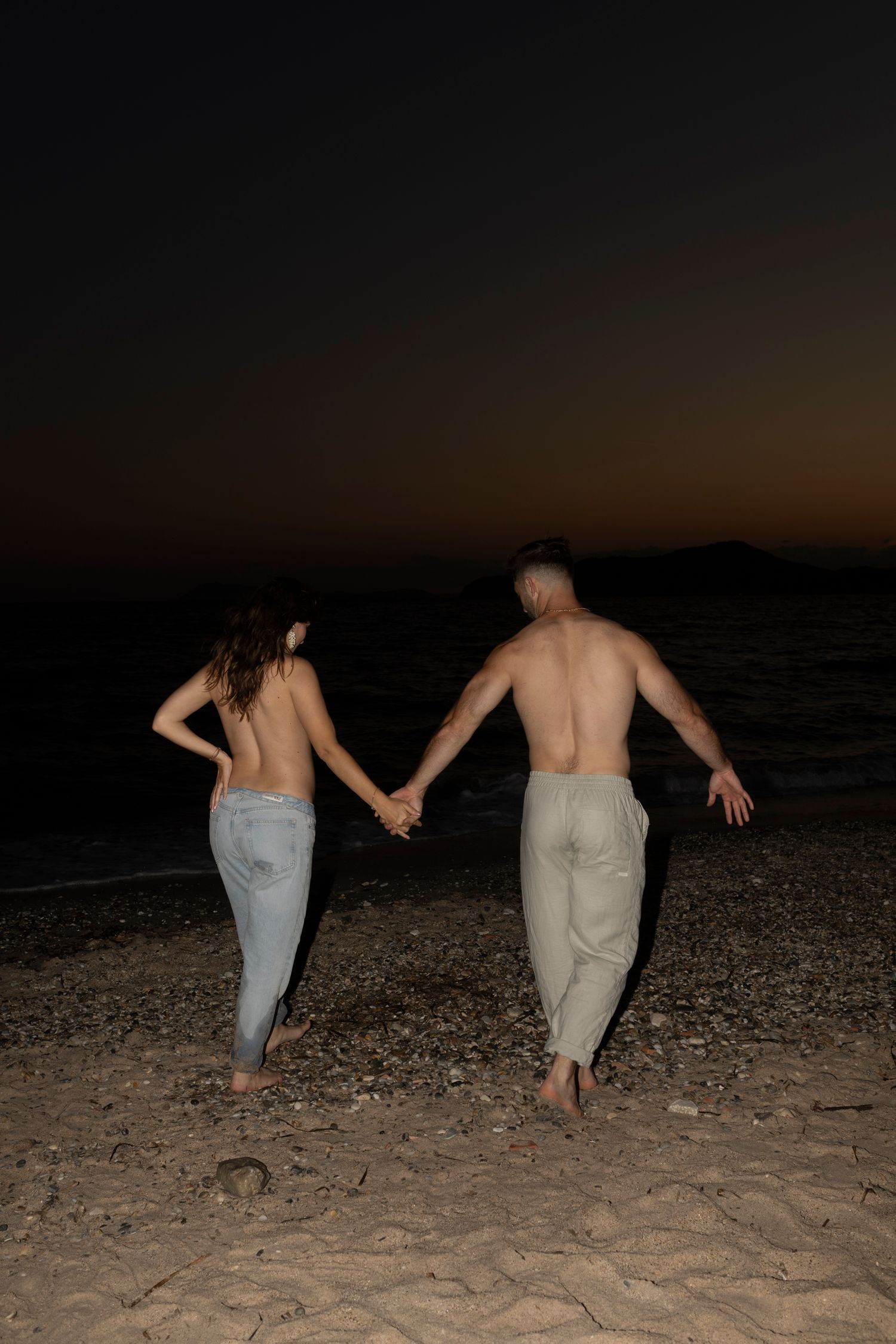 Couple holding hands while walking on beach at sunset silhouetted against dark sky.
