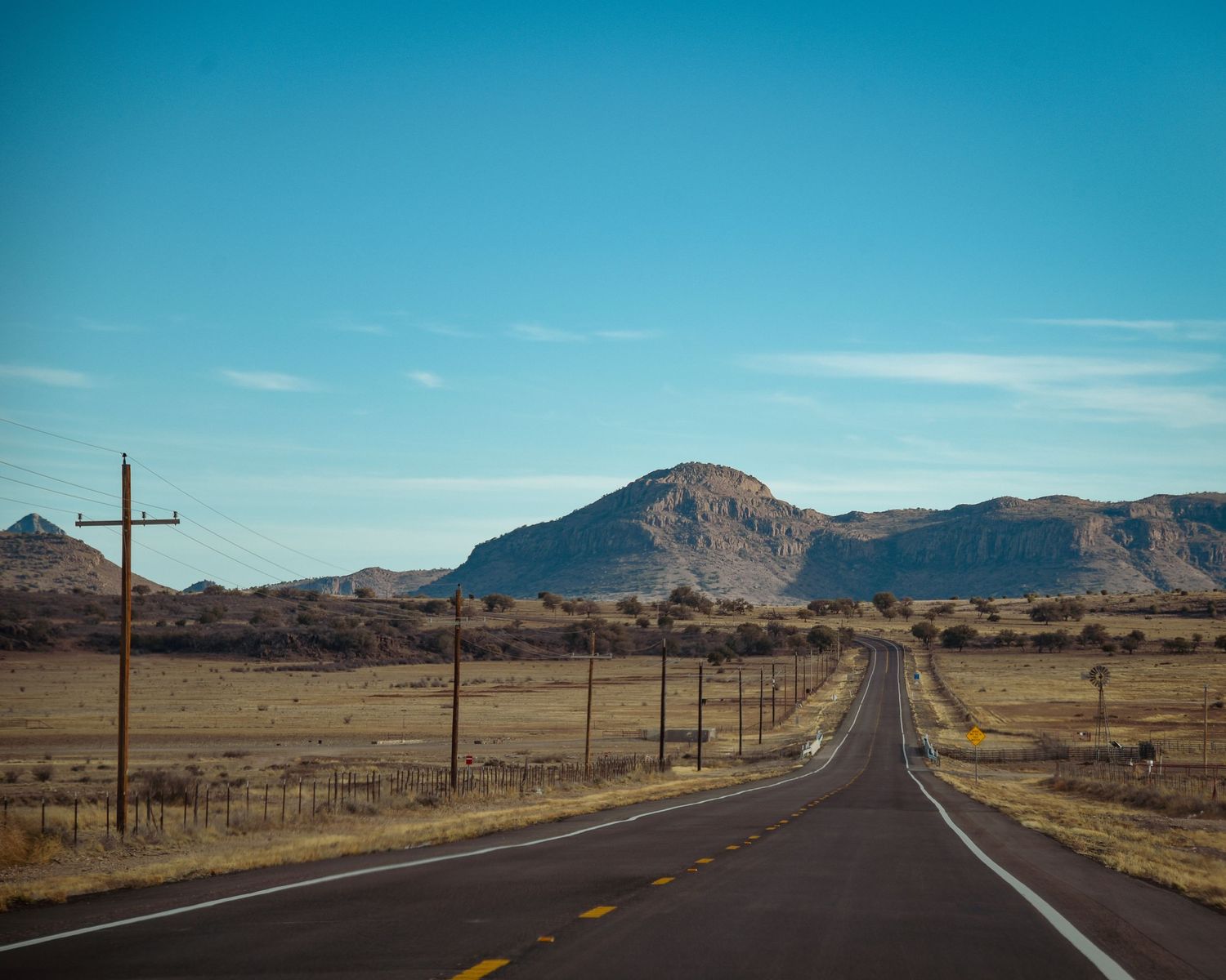 Empty desert road stretches toward mountains under bright blue sky with utility poles along the side.