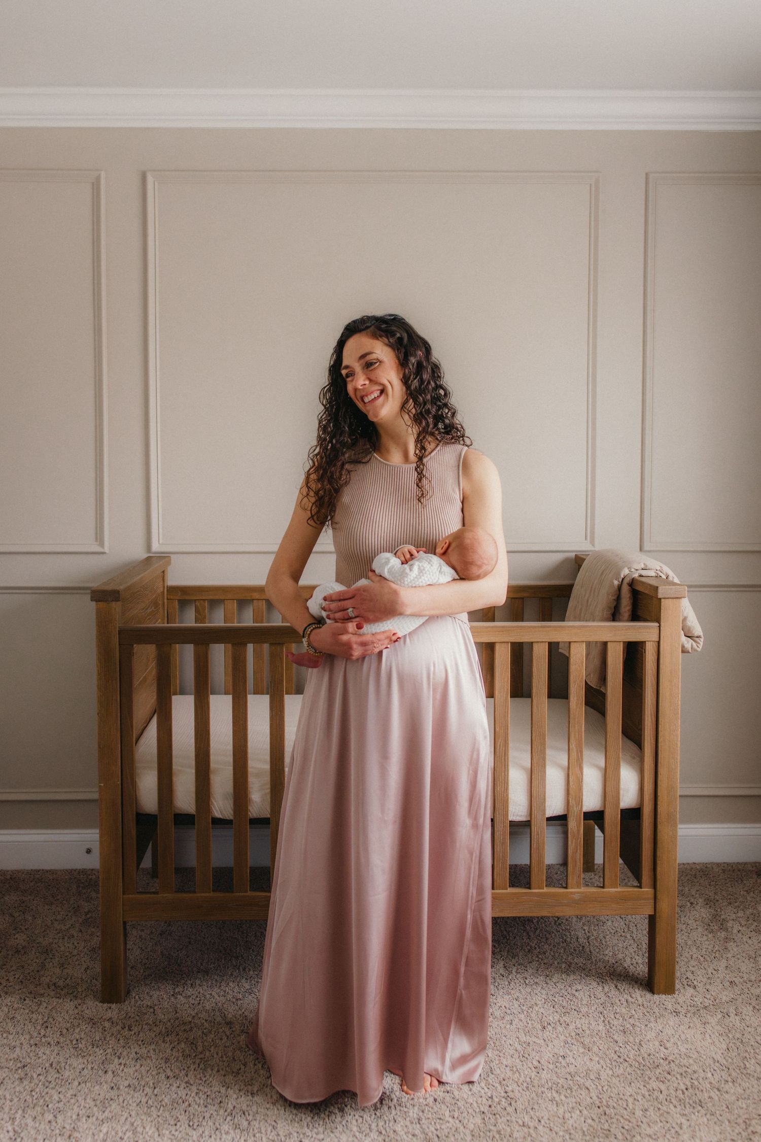 Person in a flowing pink ombre dress stands smiling next to a wooden crib in a nursery with paneled walls.